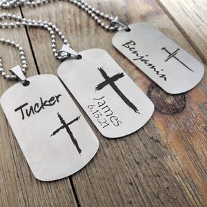 May include: Three silver dog tag necklaces with a cross design. The tags are engraved with the names "Tucker", "James 6.15.21", and "Benjamin".