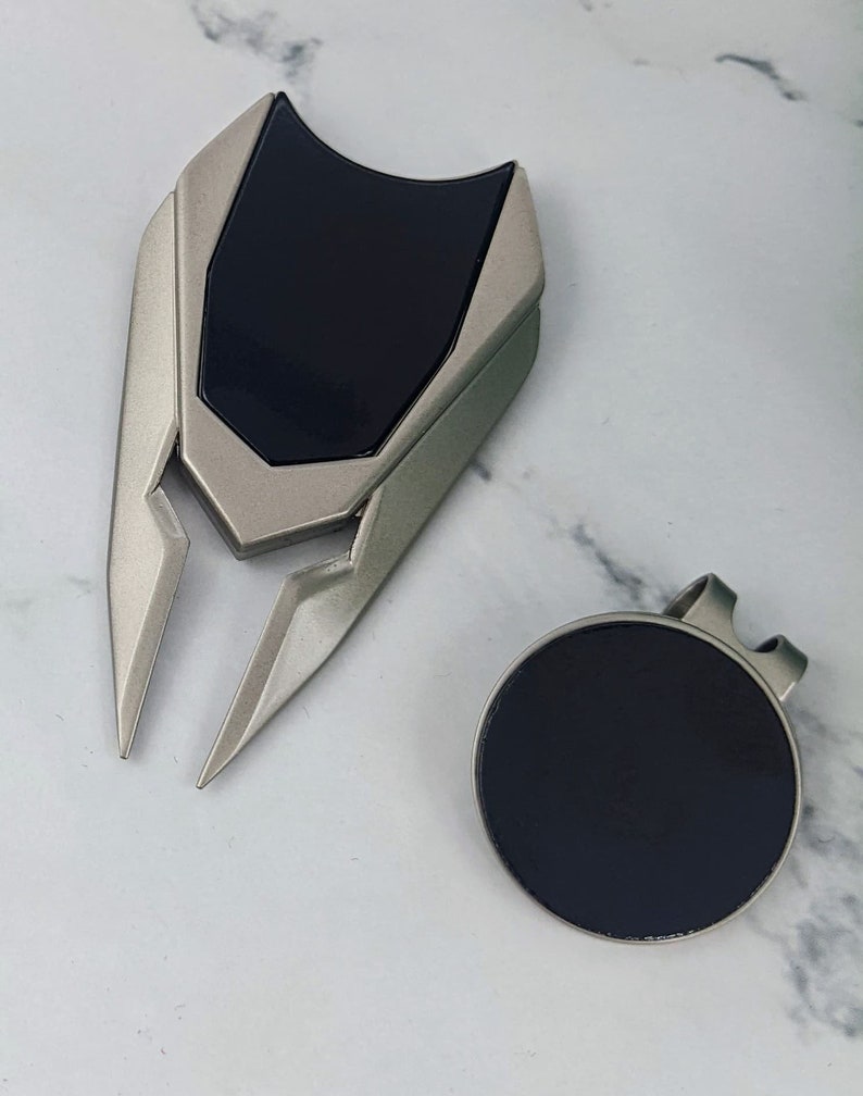 May include: Two silver golf accessories. The first is a golf ball retriever with a black rubber grip. The second is a golf ball marker with a black rubber surface.