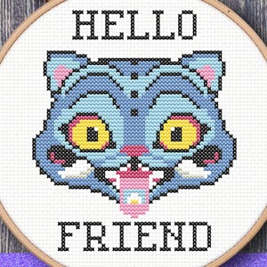 May include: A cross-stitch design in a wooden hoop featuring a blue cat face with yellow eyes and the words "HELLO FRIEND". The design is set against a backdrop of autumn leaves and a wooden surface. A digital file.
