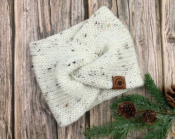 May include: White knitted headband with a brown leather tag that says "Hand Knitted with Love". The headband is on a wooden background with a pine branch.