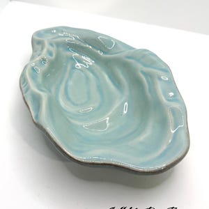 May include: A ceramic, seashell-shaped dish in a light blue hue with a glossy finish. The dish has a wavy, textured surface and a dark brown rim. The text "Call Me For Treasure" is at the bottom.