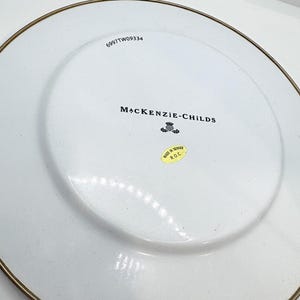 May include: White ceramic plate with a gold rim. The plate features the text "MACKENZIE-CHILDS" and a small emblem. A sticker reads "MADE IN TAIWAN R.O.C." and the number "6997TW09334" is printed on the plate.