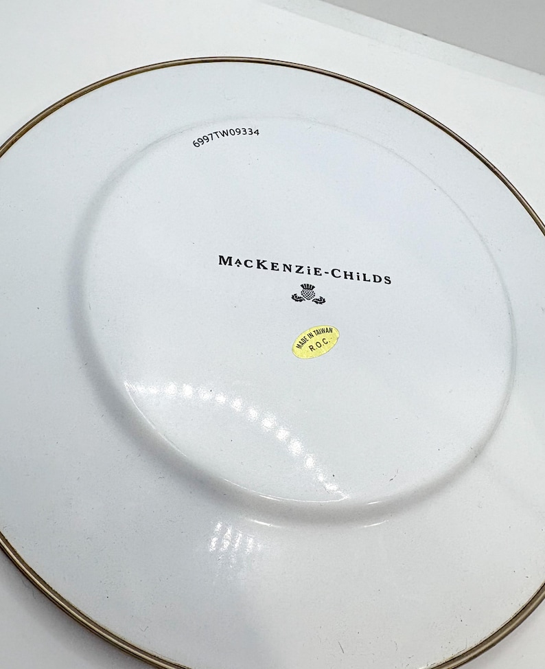 May include: White ceramic plate with a gold rim. The plate features the text "MACKENZIE-CHILDS" and a small emblem. A sticker reads "MADE IN TAIWAN R.O.C." and the number "6997TW09334" is printed on the plate.