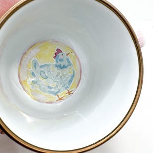 May include: White ceramic bowl with a gold rim, featuring a hand-painted light blue chicken illustration on a yellow background. The chicken has a red comb and orange beak. The bowl is likely for serving or display.