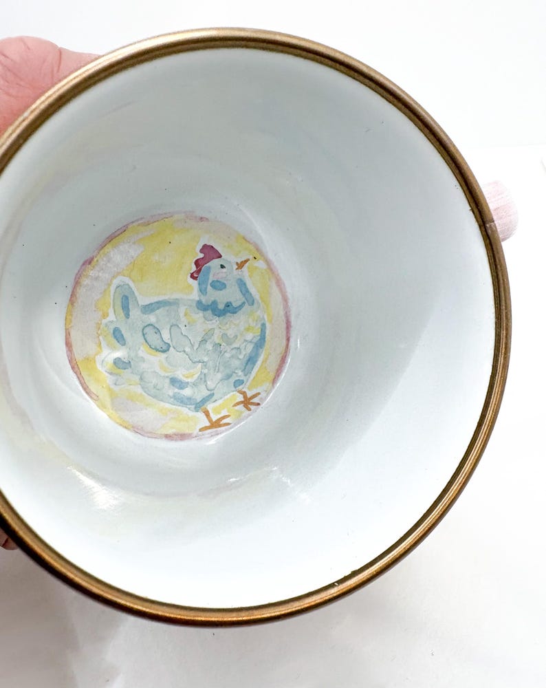 May include: White ceramic bowl with a gold rim, featuring a hand-painted light blue chicken illustration on a yellow background. The chicken has a red comb and orange beak. The bowl is likely for serving or display.