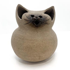 Op de afbeelding: Een keramieken kattenbeeldje met een lachend gezicht en open mond. De kat heeft grote oren en een rond lichaam. De kleur is een gespikkeld bruin met donkere accenten rond het gezicht. De tekst "Call Me For Treasure" staat onderaan.