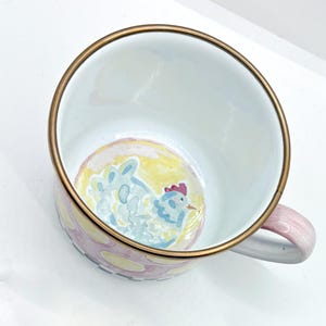 May include: A white enamel mug with a gold rim and a pink handle. The mug features a hand-painted design of a blue chicken on a yellow background. The exterior is decorated with pink and yellow polka dots.