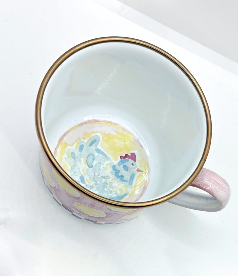 May include: A white enamel mug with a gold rim and a pink handle. The mug features a hand-painted design of a blue chicken on a yellow background. The exterior is decorated with pink and yellow polka dots.