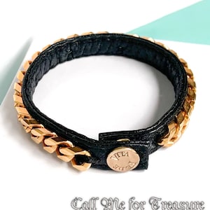 May include: A black leather bracelet with a gold chain detail and a gold button closure. The button has the text "VITA FE" engraved on it.