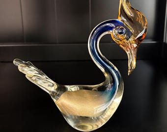 Peut inclure: Une figurine d'oiseau en verre avec un corps transparent et une base à paillettes dorées, un cou bleu profond et une tête ainsi qu'un bec de couleur ambre. L'oiseau est représenté dans une posture penchée vers le bas avec une queue pointue.