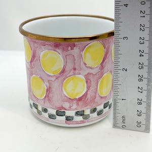 May include: A cylindrical ceramic mug with a pink watercolor background and yellow polka dots. The rim is trimmed with gold, and the base features a black and white checkered pattern. The mug is approximately 3 inches tall.