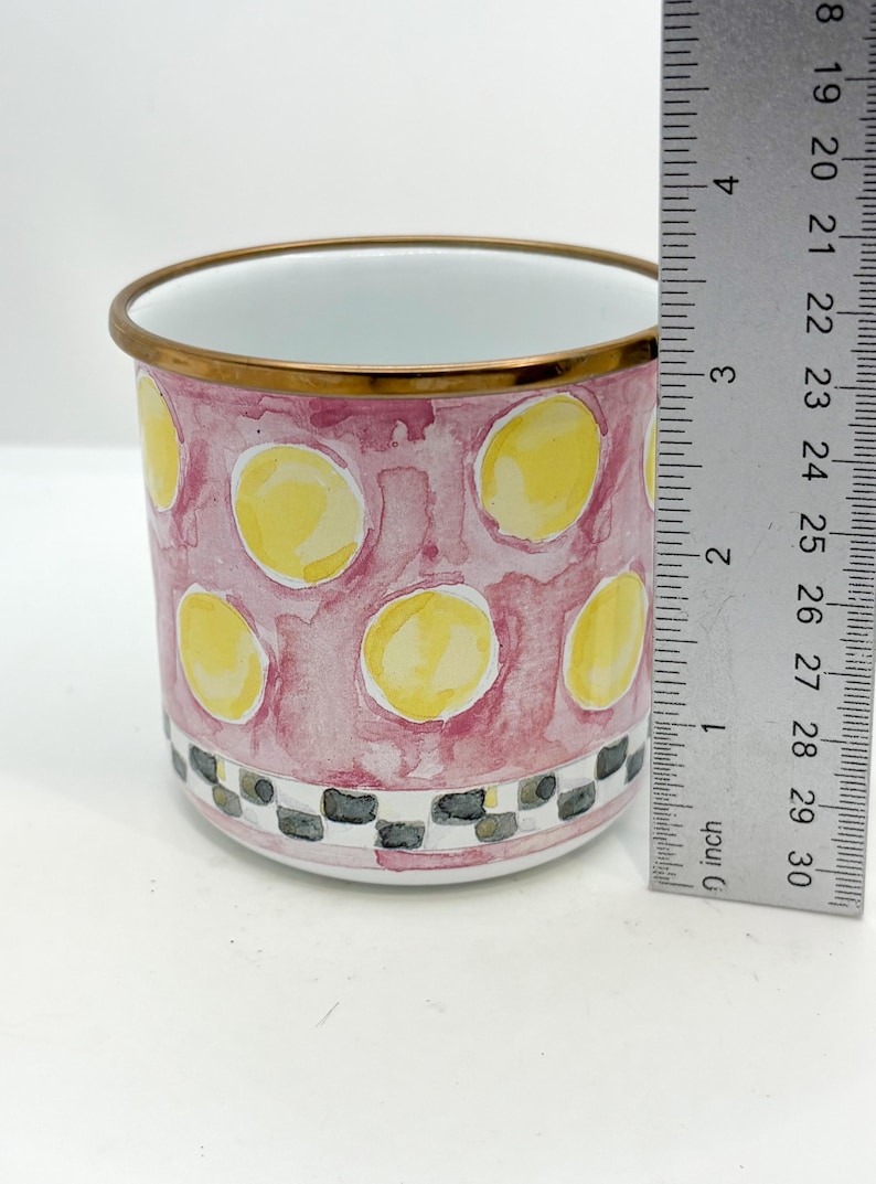 May include: A cylindrical ceramic mug with a pink watercolor background and yellow polka dots. The rim is trimmed with gold, and the base features a black and white checkered pattern. The mug is approximately 3 inches tall.