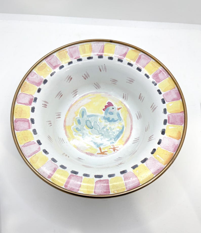 May include: A decorative ceramic bowl with a hand-painted chicken design. The bowl features a yellow and pink checkered border with a gold rim. The chicken is light blue with a red comb, set against a yellow background.