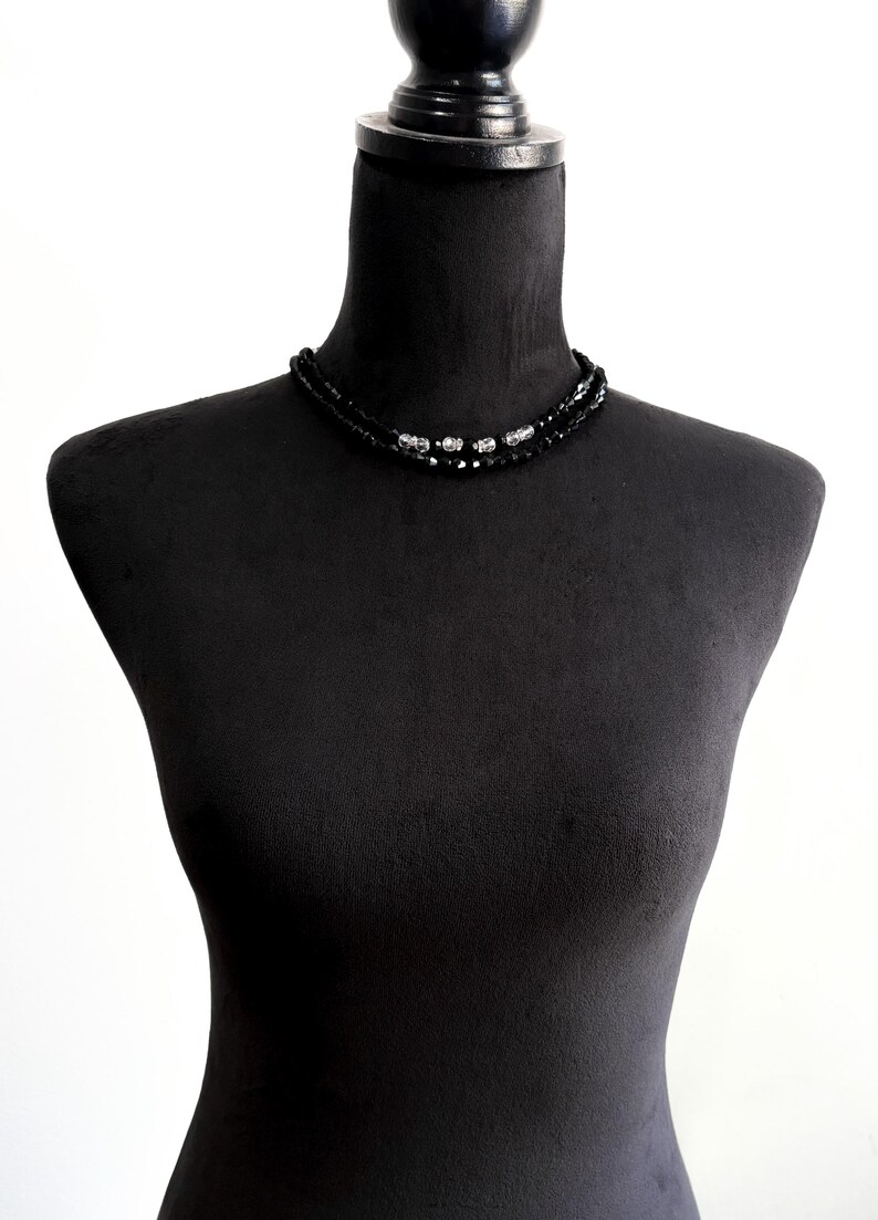 May include: A black beaded necklace with silver accents, displayed on a black mannequin. The necklace features two strands of small, dark beads and several larger, decorative silver beads.