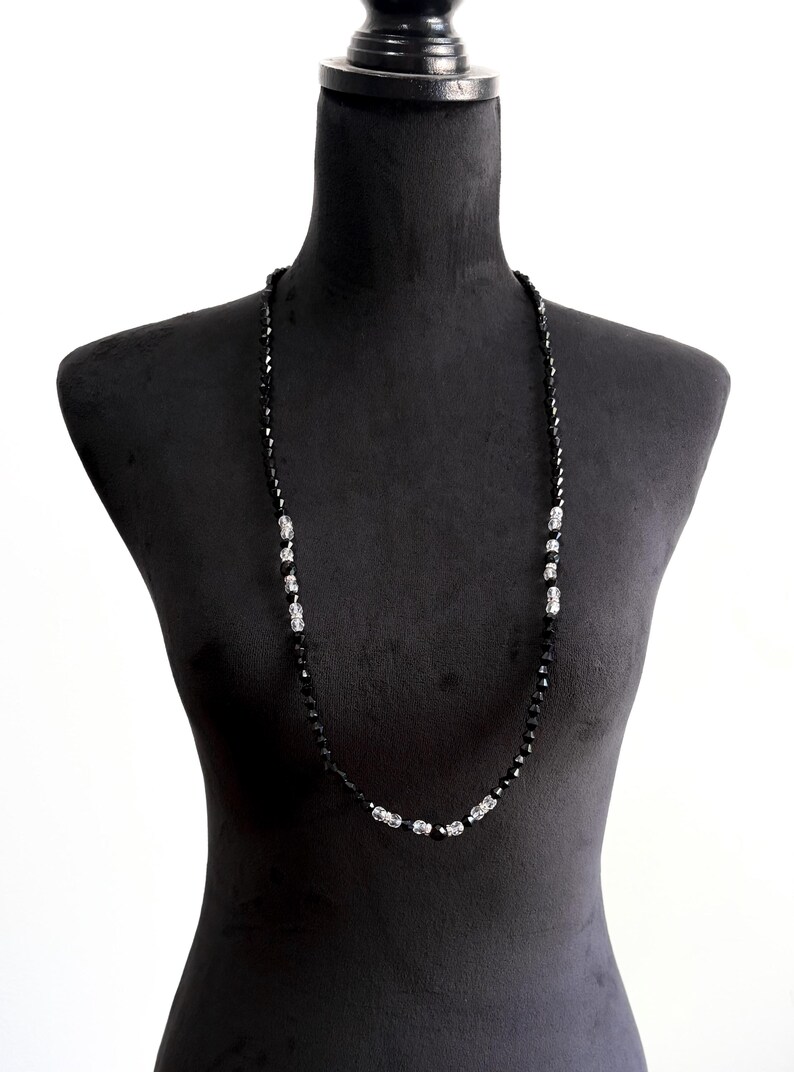 May include: A long black beaded necklace with silver accents, displayed on a black mannequin. The necklace features small, faceted black beads with sections of silver-coloured beads.