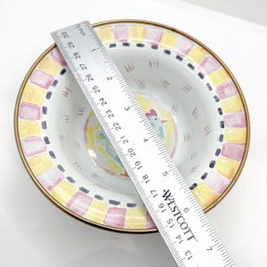 May include: A decorative ceramic bowl with a painted rooster design in the center. The bowl has a gold rim and a border of yellow and pink squares. The bowl measures approximately 10 inches in diameter.