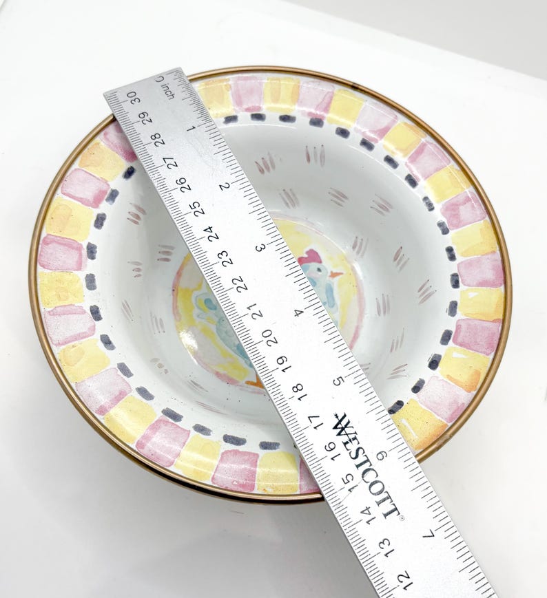 May include: A decorative ceramic bowl with a painted rooster design in the center. The bowl has a gold rim and a border of yellow and pink squares. The bowl measures approximately 10 inches in diameter.