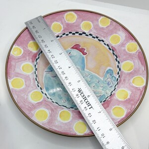 May include: A decorative ceramic plate with a hand-painted design. The plate features a light blue chicken in the center, surrounded by a pink border with yellow polka dots. The plate is approximately 12 inches in diameter.