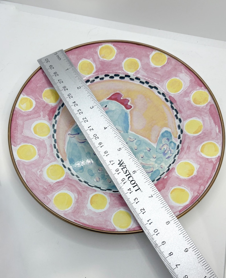 May include: A decorative ceramic plate with a hand-painted design. The plate features a light blue chicken in the center, surrounded by a pink border with yellow polka dots. The plate is approximately 12 inches in diameter.