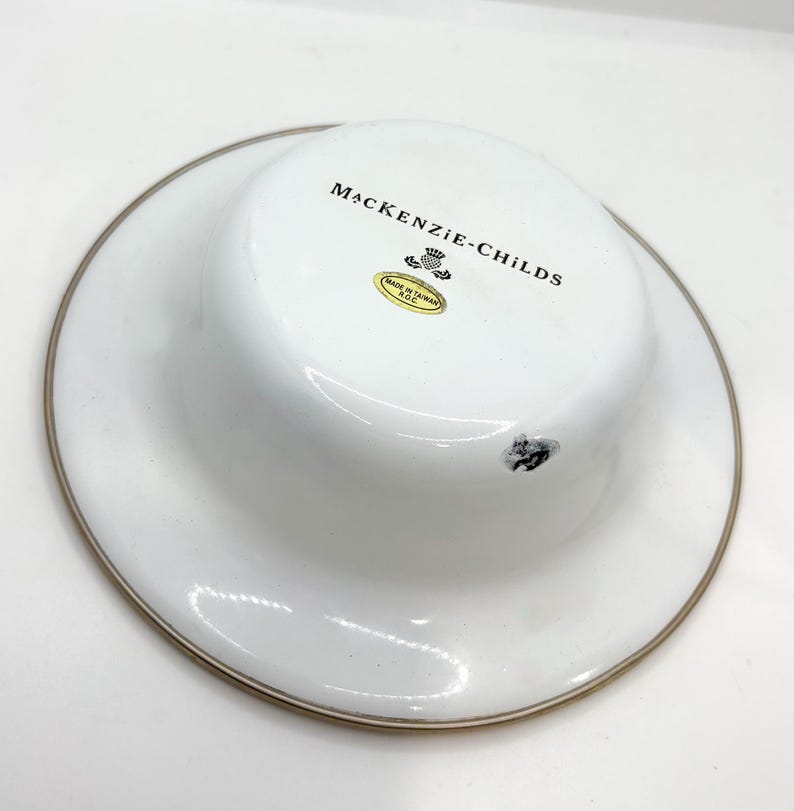 May include: White ceramic dish with a gold rim. The dish has a raised center and features the text "MacKenzie-Childs" in black. A small sticker is visible, along with a small black smudge.