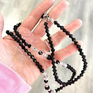 May include: A black beaded necklace with clear crystal detailing. The necklace is made up of black, faceted beads interspersed with clear crystal beads. It is secured with a silver clasp. The necklace is shown against a neutral backdrop.