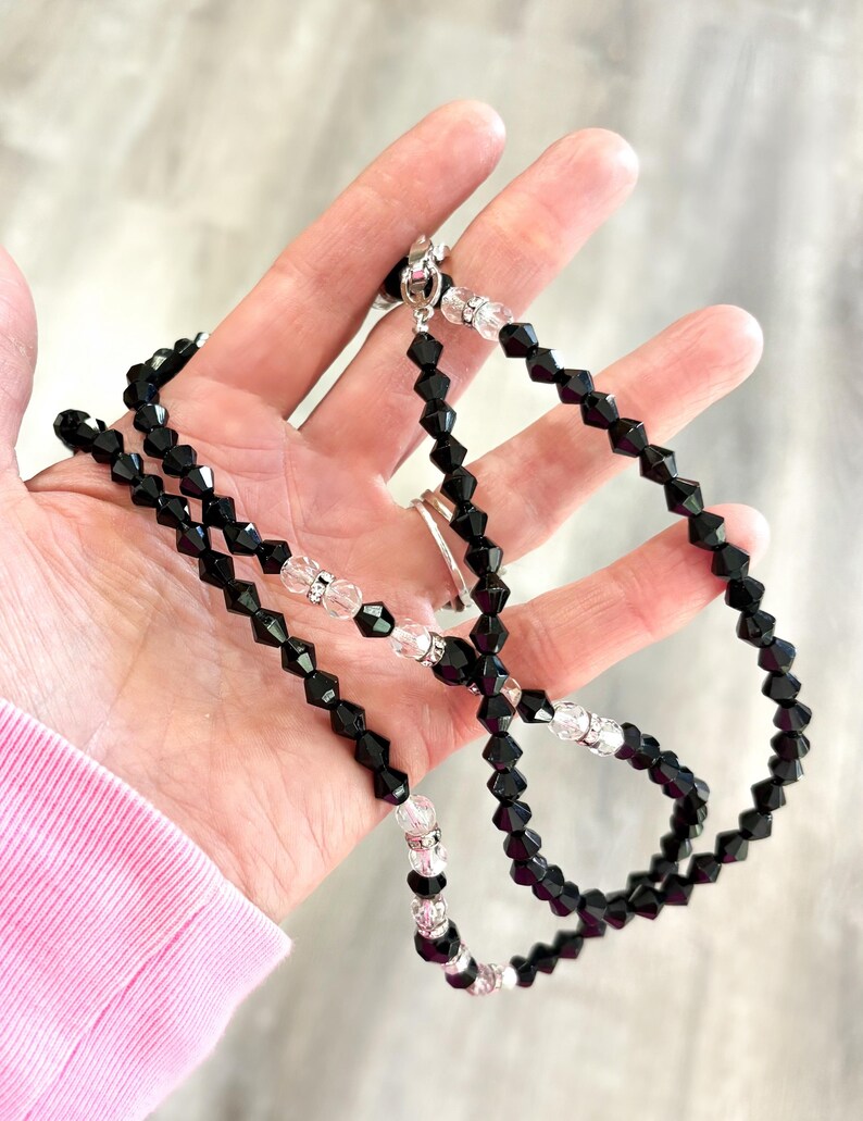 May include: A black beaded necklace with clear crystal detailing. The necklace is made up of black, faceted beads interspersed with clear crystal beads. It is secured with a silver clasp. The necklace is shown against a neutral backdrop.