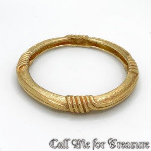 Puede incluir: Un brazalete dorado con una superficie texturizada y detalles acanalados. El brazalete tiene un diseño circular con una forma ligeramente ovalada. El texto "Call Me for Treasure" está en la parte inferior.