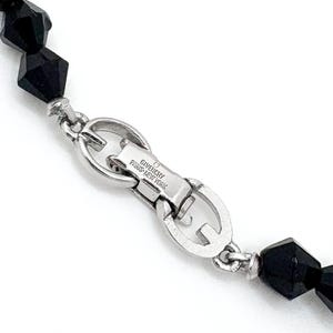 May include: Close-up of a black beaded necklace with a silver clasp. The clasp is engraved with "GIVENCHY PARIS-NEW YORK". The beads are faceted and have a geometric shape. The necklace is on a white background.