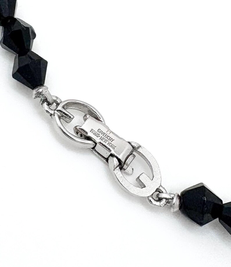 May include: Close-up of a black beaded necklace with a silver clasp. The clasp is engraved with "GIVENCHY PARIS-NEW YORK". The beads are faceted and have a geometric shape. The necklace is on a white background.