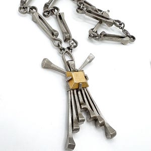 May include: A silver-toned metal necklace with a unique design. The necklace features a series of rectangular links and a pendant with a gold-colored square center and radiating metal elements. The text "Call Me For Treasure" is visible at the bottom.
