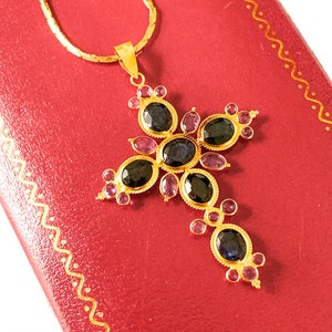 May include: A gold chain necklace with a pendant in the shape of a cross. The cross is adorned with dark blue and pink gemstones.