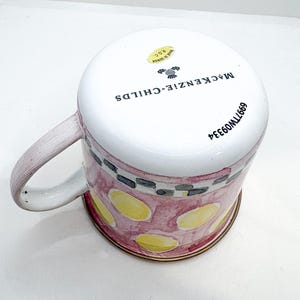 May include: A ceramic mug with a white top and a pink and yellow patterned body. The mug has a handle and a gold-colored rim. The text "MACKENZIE-CHILDS" is printed on the side of the mug.