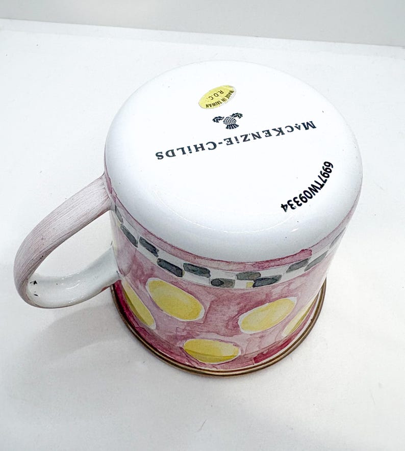 May include: A ceramic mug with a white top and a pink and yellow patterned body. The mug has a handle and a gold-colored rim. The text "MACKENZIE-CHILDS" is printed on the side of the mug.
