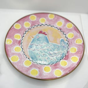 May include: A decorative plate with a hand-painted design. The plate features a light blue chicken in the center, surrounded by a yellow and orange background. The rim is pink with yellow polka dots and a gold-colored edge.