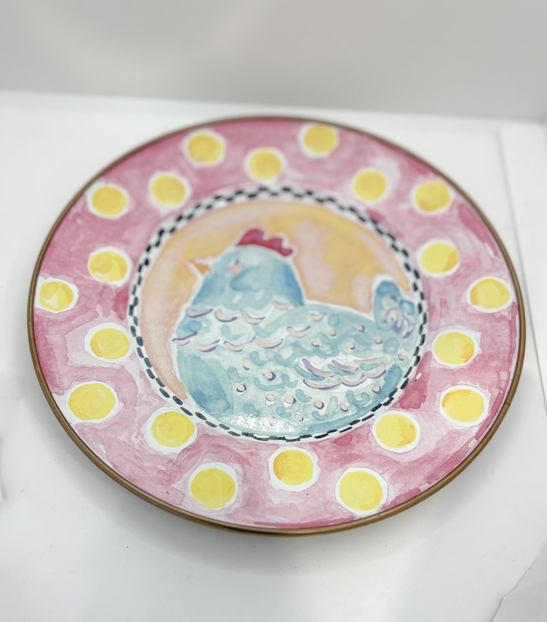 May include: A decorative plate with a hand-painted design. The plate features a light blue chicken in the center, surrounded by a yellow and orange background. The rim is pink with yellow polka dots and a gold-colored edge.