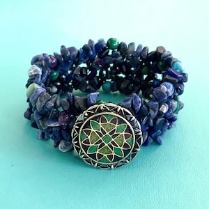 May include: A multi-strand bracelet made of blue, green and black gemstone chips. The bracelet has a silver clasp with a decorative green and blue design.