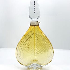May include: A heart-shaped amber glass perfume bottle with a clear glass stopper featuring the word CHAMADE. The base of the bottle is clear glass and features the embossed text GUERLAIN. The bottle has a ribbed texture throughout the body.