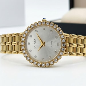 May include: A gold-tone quartz wristwatch with a round silver dial featuring the text LAROSA and QUARTZ. The bezel is encrusted with clear stones, and the dial includes small stone hour markers. It has a gold-tone metal link bracelet.