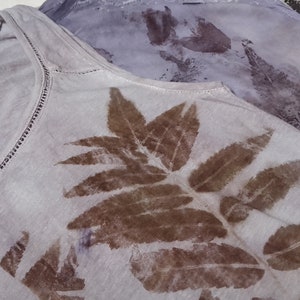 May include: A white tank top with a brown leaf design printed on the front. The leaves are arranged in a symmetrical pattern.