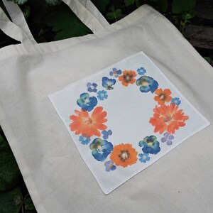 May include: A white canvas tote bag with a floral design on a pocket. The design features orange and blue flowers in a circular pattern.