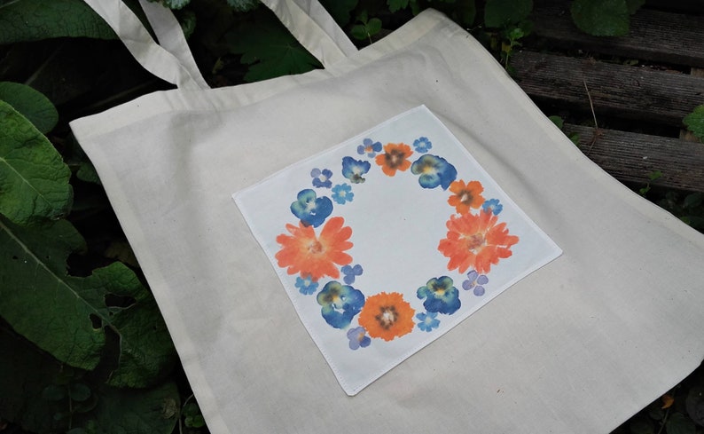 May include: A white canvas tote bag with a floral design on a pocket. The design features orange and blue flowers in a circular pattern.