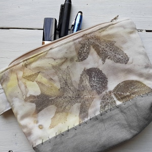 May include: A white zippered pouch with a gray bottom. The pouch is decorated with a leaf pattern in shades of brown, yellow, and green.