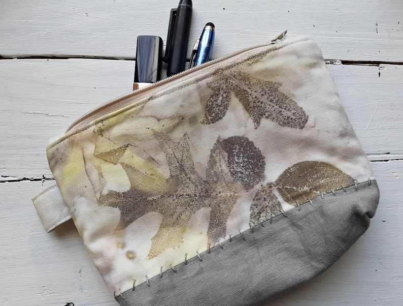 May include: A white zippered pouch with a gray bottom. The pouch is decorated with a leaf pattern in shades of brown, yellow, and green.