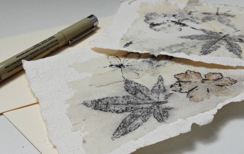 May include: Two pieces of white paper with delicate, pressed leaves and flowers. The leaves are in shades of brown and gray, and the flowers are white. The paper has a textured surface.