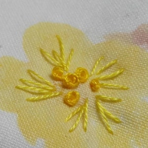May include: Close-up of a yellow embroidered flower design on a white fabric background.