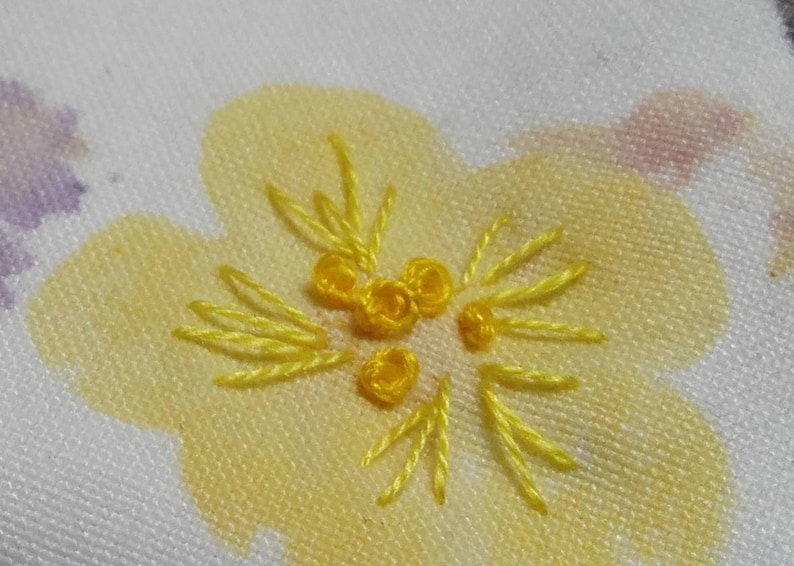 May include: Close-up of a yellow embroidered flower design on a white fabric background.