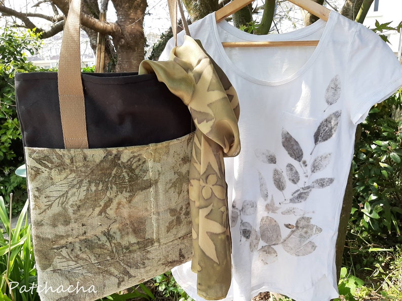 May include: A black tote bag with a brown strap and a green and brown floral print on the front. A white t-shirt with a green and brown leaf print is hanging on a wooden hanger. A green and brown floral print scarf is draped over the hanger.