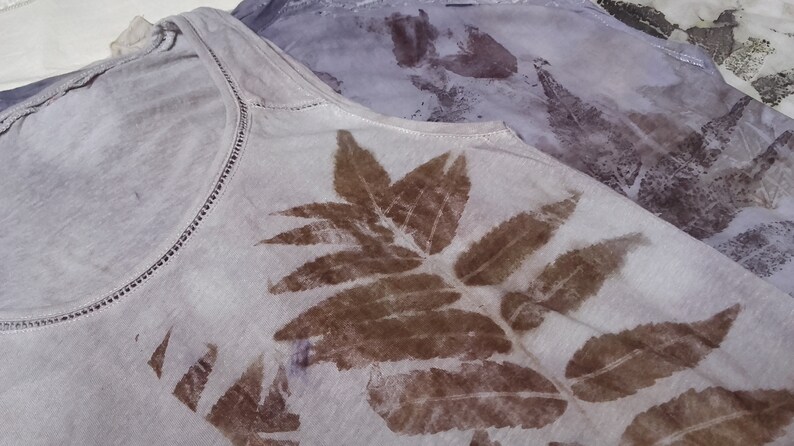 May include: A white tank top with a brown leaf design printed on the front. The leaves are arranged in a symmetrical pattern.