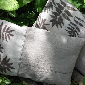 May include: Two decorative throw pillows with a fern leaf design printed on a light gray linen fabric. The pillows are sitting on a wooden surface.