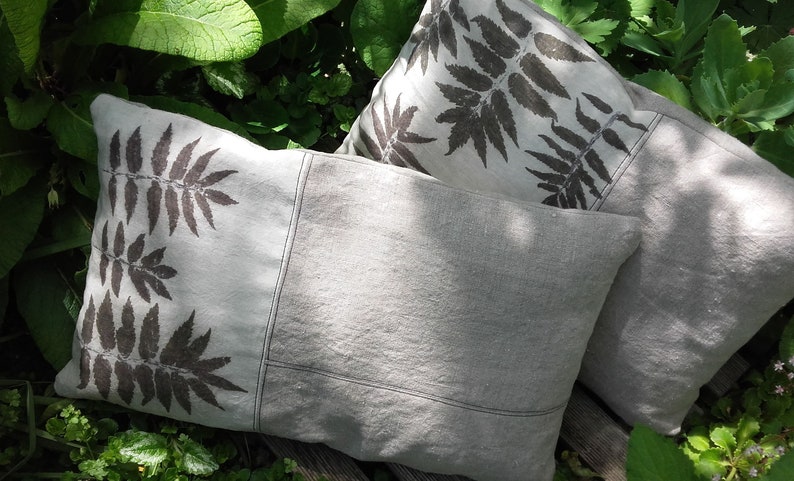 May include: Two decorative throw pillows with a fern leaf design printed on a light gray linen fabric. The pillows are sitting on a wooden surface.